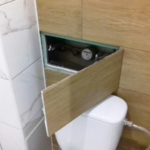 Panel de acceso oculto en la pared del baño con ducha y aseo alicatado. Puerta de trampilla a medida. Tapa de inspección de servicios invisible. Mecanismo de cierre por presión.