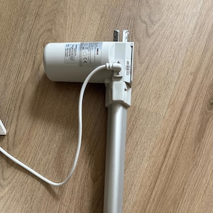 Può includere: Un attuatore lineare color argento con un alloggiamento motore cilindrico bianco e un cavo di alimentazione bianco. L'attuatore è su una superficie di legno. L'alloggiamento del motore ha un'etichetta con testo.