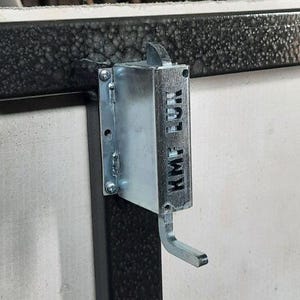 May include: Close-up of a metal latch with the text "KMF LUN" on a black metal frame. The frame has a grid-like design and is attached to a white surface. The latch is silver and appears to be functional.