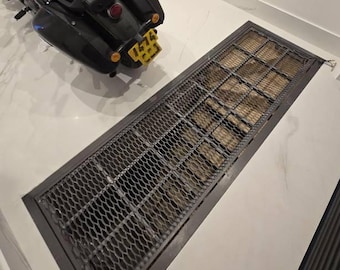 May include: A black motorcycle with a yellow license plate parked next to a rectangular floor grate featuring a dark metal frame and a grid pattern, set into a white marble-patterned floor surface with a black ribbed wall section nearby.