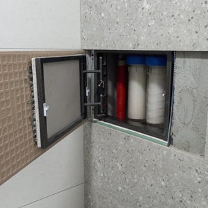 Może przedstawiać: Wbudowany system filtracji wody z otwartym panelem dostępowym. System zawiera trzy cylindryczne filtry, jeden czerwony i dwa białe, z niebieskimi nakrętkami. Panel jest otwarty, odsłaniając filtry w zagłębieniu w nakrapianej szarej ścianie.