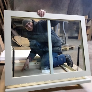 Puede incluir: Un marco de ventana blanco con un panel de vidrio transparente. La ventana está abierta y una persona está de pie detrás. La persona lleva una chaqueta azul y un sombrero negro.