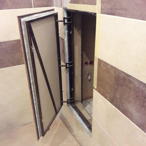 Könnte beinhalten: Eine offene, rechteckige Zugangsklappe mit Metallrahmen und hellem Innenraum. Die Klappe ist in einer gefliesten Wand mit abwechselnd braunen und beigen Fliesen installiert. Der Rahmen ist dunkel.