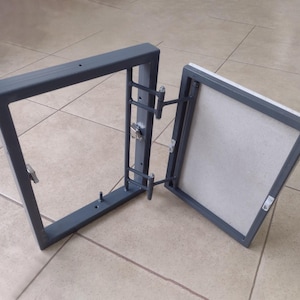 Può includere: Un pannello di accesso in metallo grigio, aperto per rivelare un interno di colore chiaro. Il pannello è rettangolare con una porta a cerniera e un fermo. Il telaio è grigio scuro, su un pavimento piastrellato.