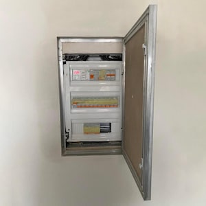 Puede incluir: Un panel eléctrico blanco con una puerta plateada está montado en una pared blanca. El panel tiene varios interruptores y una pantalla digital que muestra "24H".