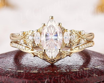 May include: A gold-colored engagement ring set featuring a marquise-cut center stone and smaller accent stones. The band has intricate floral detailing. The ring is displayed on a red jewelry box.