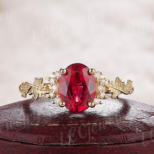 Könnte beinhalten: Ein goldener Ring mit einem ovalen Rubin-Edelstein, der von kleineren Diamanten umgeben ist. Der Ring hat ein zartes Blattdesign auf jeder Seite des Rubins.