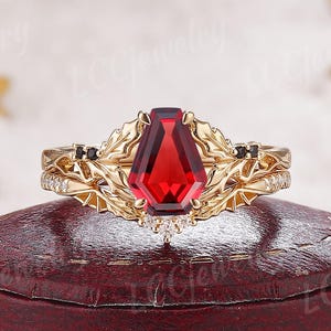 May include: A gold ring set featuring a large, rectangular, deep red gemstone. The ring is adorned with intricate leaf-like designs and small black stones. The ring is displayed on a red jewelry box, set against a snowy background.