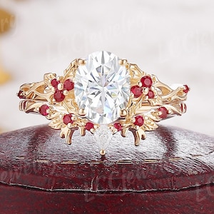 May include: A gold-colored ring with an oval, clear center stone and small red accent stones. The ring has a floral design with intricate details. The ring is displayed on a dark red surface.