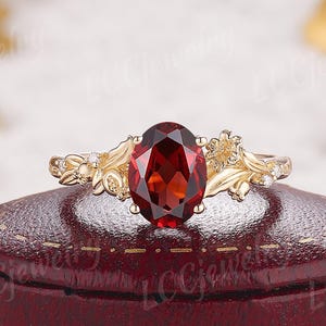 May include: A gold ring featuring a central oval-cut deep red garnet gemstone. The band is designed with intricate metal leaf and vine motifs on both sides of the stone, accented with small, clear, sparkling round-cut stones.