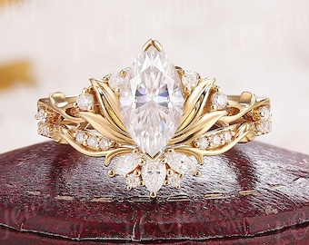 May include: A gold engagement ring with a marquise-cut center stone and a floral design. The ring features smaller accent stones and is set on a red jewelry box. The ring's design includes leaf and petal details.