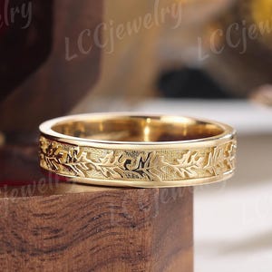 May include: A gold wedding band with a leaf design. The band is sitting on a wooden box.