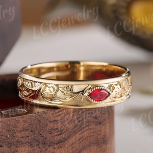 Puede incluir: Un anillo de banda de oro con una piedra roja engastada en el centro. La banda está decorada con un intrincado diseño floral.