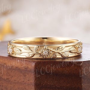 Puede incluir: Una banda de boda de oro con un delicado diseño floral y pequeños diamantes. La banda está sobre una superficie de madera.