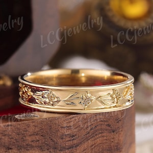 May include: A gold-colored ring with a floral design. The ring features a repeating pattern of flowers and leaves around the band. The ring is displayed on a wooden surface.
