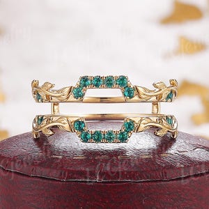 May include: A gold-colored ring guard with emerald-colored gemstones. The ring guard has a floral design on the sides and is set on a red jewelry box. The gemstones are set in a geometric pattern.