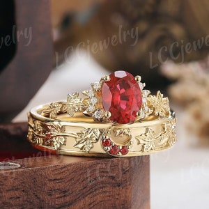 Könnte beinhalten: Ein Set aus zwei goldenen Ringen mit einem roten Edelstein und Blattmotiven. Der Verlobungsring hat einen großen ovalen roten Edelstein, der von kleineren Diamanten umgeben ist. Der Ehering hat ein Blattmotiv und einen Halbmond mit einem kleinen roten Edelstein.