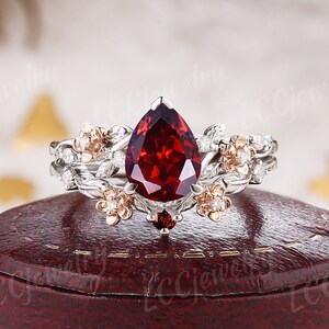 May include: A pear-shaped red garnet engagement ring set in white gold with a floral design and smaller diamonds. The ring is on a red velvet box.