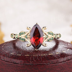 May include: A gold ring featuring a marquise-shaped red gemstone surrounded by smaller green gemstones. The ring is displayed against a white backdrop.