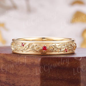 May include: A gold band ring featuring an intricate relief pattern of sculpted leaves and flowers. The metal surface is polished and includes three small, round, vibrant red gemstones set at intervals within the floral design.