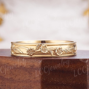 Könnte beinhalten: Ein goldfarbener Ring mit floralem Design. Der Ring hat geprägte Blumen und Blätter um das Band. Der Ring wird auf einer dunklen Holzoberfläche präsentiert.