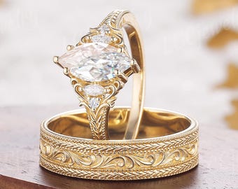 May include: A yellow gold engagement ring set featuring a marquise-cut center stone flanked by smaller accent stones on a filigree band, paired with a matching yellow gold wedding band that displays an intricate engraved scrollwork pattern.