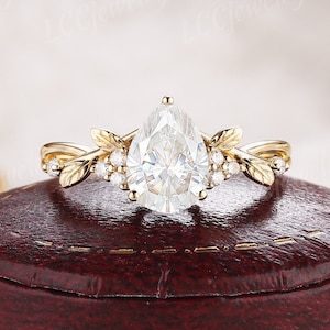 May include: A pear-shaped diamond engagement ring set in a gold band with delicate leaf accents. The ring is displayed in a red velvet box.