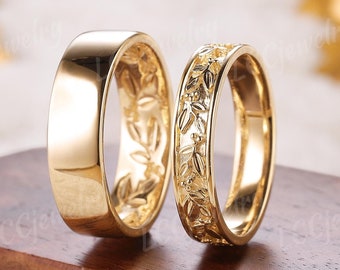 Puede incluir: Dos anillos de boda dorados. Uno es liso y pulido, mientras que el otro presenta un detallado diseño de hojas. Los anillos se exhiben sobre una superficie de madera oscura, resaltando su acabado brillante y reflectante. Los anillos son un símbolo de compromiso.