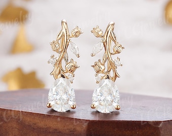 May include: A pair of gold-colored earrings with a floral design. Each earring features a pear-shaped, clear gemstone at the bottom, with delicate branches adorned with smaller clear stones and floral accents. The earrings are displayed on a wooden surface.