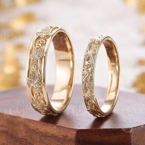 May include: Two gold wedding rings with a detailed leaf pattern. The rings are displayed on a wooden surface, showcasing their intricate design. The rings are a matching set, with one slightly larger than the other.