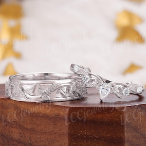 Puede incluir: Dos anillos de plata. Uno presenta un diseño de hojas detallado. El segundo tiene un diseño curvo con pequeñas piedras claras y detalles de hojas. Los anillos se exhiben sobre un bloque de madera.