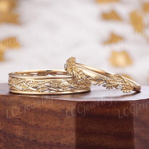 Puede incluir: Dos anillos de boda de oro. Uno tiene una banda con un patrón de hojas detallado. El otro tiene un diseño de hojas y diamantes. Los anillos se exhiben sobre una superficie de madera.