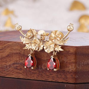 May include: A pair of gold-colored floral earrings with teardrop-shaped, deep red gemstones. The earrings feature intricate flower and leaf details, with small, clear accent stones. The earrings are displayed on a wooden surface.