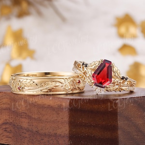 May include: A gold wedding ring set. The band features an engraved leaf design with small red accents. The engagement ring has a large, red, pear-shaped gemstone, black accents, and small white gemstones.