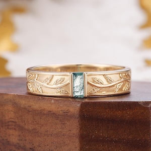 May include: A gold-colored ring with a rectangular, light-green gemstone. The band features a leaf and vine pattern. The ring is displayed on a dark wooden surface.