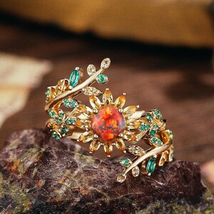 May include: A gold-toned ring featuring a central, fiery red gemstone surrounded by smaller yellow stones, resembling a flower. Green gemstones and delicate leaf-like details adorn the band, creating a nature-inspired design. The ring is set on a textured, dark-colored stone.