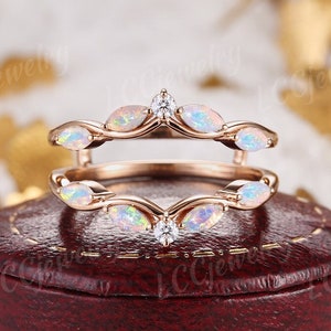 以下が含まれることがあります： オパールとダイヤモンドのアクセントが付いたローズゴールドのリングガード。リングは、楕円形のオパールと小さなダイヤモンドを備えた葉のようなデザインが特徴です。リングは赤いジュエリーボックスに展示されています。