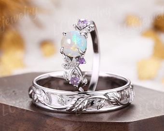 Könnte beinhalten: Ein Set aus zwei silbernen Ringen mit einem floralen Design. Der Verlobungsring hat einen großen, ovalen Opal-Edelstein mit kleineren violetten Edelsteinen auf der Band. Der Ehering hat ein passendes florales Design.