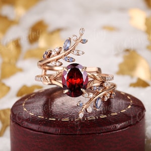 Puede incluir: Un anillo de oro rosa con una gran piedra roja ovalada rodeada de pequeñas piedras blancas. El anillo presenta un diseño delicado de ramas entrelazadas con hojas y pequeñas piedras azules.