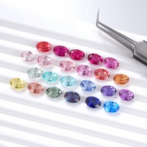 May include: A collection of oval-cut gemstones in a variety of colors, including red, pink, blue, and yellow. The gemstones are arranged on a white surface, with a pair of tweezers in the background. The gemstones are likely for jewelry making.