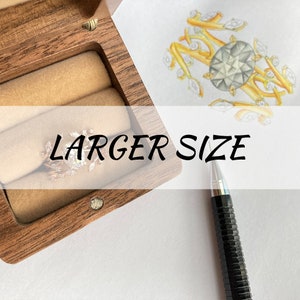 Puede incluir: Una caja de madera para anillos con un anillo dentro. El anillo es un diseño delicado con una piedra central grande y piedras más pequeñas alrededor. La caja está abierta y el anillo es visible. El texto "LARGER SIZE" está escrito en la imagen.