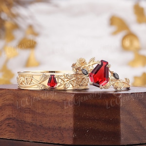 Puede incluir: Dos anillos de oro con intrincados diseños florales y de hojas. Un anillo presenta una gran gema roja con corte ataúd y dos gemas negras. El otro anillo tiene una gema roja más pequeña. Los anillos se exhiben sobre una superficie de madera.