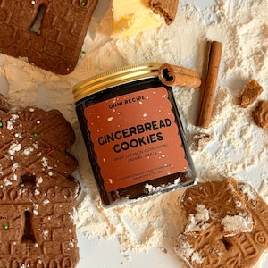 Op de afbeelding: Een glazen pot met een gouden deksel en een etiket met de tekst ONNI RECIPE GINGERBREAD COOKIES, GINGER CINNAMON CLOVE NUTMEG CARAMEL VANILLA, RAPESEED BLEND HAND POURED IN NL, ONNIRECIPE.ETSY.COM, omringd door peperkoekjes, een kaneelstokje en boter.