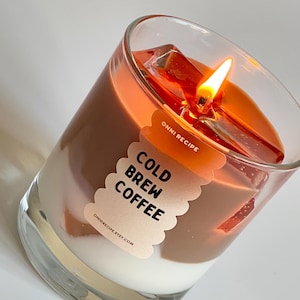 Op de afbeelding: Een glazen kaars met drie lagen was, die lijkt op een koude koffie-drank. De bovenste laag is oranje, de middelste laag is bruin en de onderste laag is wit. De kaars is aangestoken en heeft een label met de tekst "COLD BREW COFFEE" met de website "ONNIRECIPE.ETSY.COM".