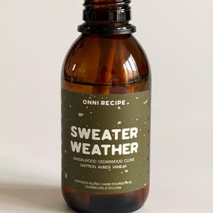 Può includere: Una bottiglia di vetro marrone con tappo nero e bastoncini di legno. L'etichetta recita "SWEATER WEATHER" con note olfattive di legno di sandalo, legno di cedro, chiodi di garofano, zafferano, ambra e vaniglia. Miscela di colza.
