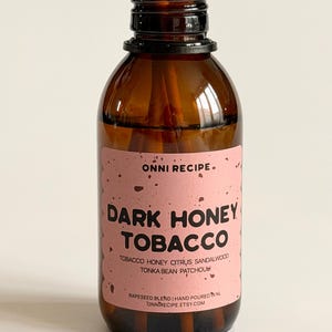 Peut inclure: Une bouteille en verre marron avec un bouchon noir, remplie de roseaux pour la diffusion de parfum. L'étiquette indique "DARK HONEY TOBACCO" avec des notes olfactives. Produit de parfum d'intérieur.