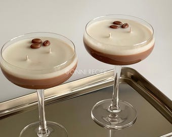 May include: Two glass coupe glasses filled with layered brown and white wax, resembling coffee cocktails. Each candle features two wicks and is topped with two brown coffee bean-shaped wax decorations. The text ONNI RECIPE is visible.