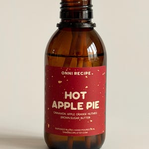 Peut inclure: Un flacon diffuseur en verre brun avec une étiquette rouge indiquant "HOT APPLE PIE". L'étiquette énumère les ingrédients comme la cannelle et la muscade. Des bâtonnets de roseau sont insérés dans le flacon.