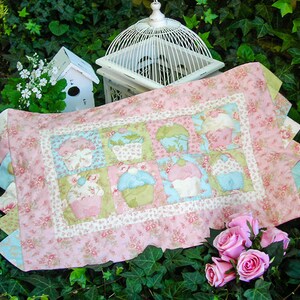 May include: A pink and green patchwork quilt with a floral pattern and a design of cupcakes. The quilt is on a bed of green leaves.