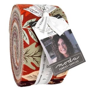 May include: A tightly wound roll of fabric strips featuring a pattern of autumn leaves in shades of orange, red, and cream. A white ribbon with a printed ruler design secures the roll, attached to a tag with the text Moda Fabrics + Supplies.